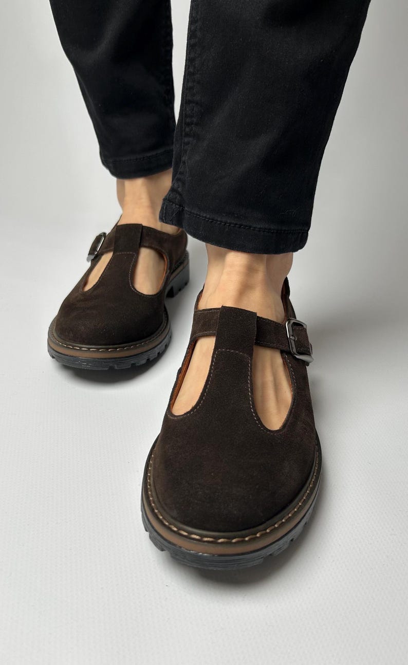 Puede incluir: Primer plano de zapatos marrones de ante con correa en T y cierre de hebilla. Los zapatos tienen punta redonda y suela gruesa y texturizada. El fondo es una superficie blanca lisa.