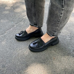May include: Black leather loafers with a tassel detail and a chunky platform sole. The shoes are worn with dark gray jeans.