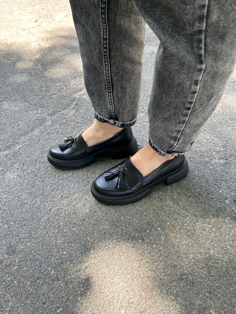 May include: Black leather loafers with a tassel detail and a chunky platform sole. The shoes are worn with dark gray jeans.