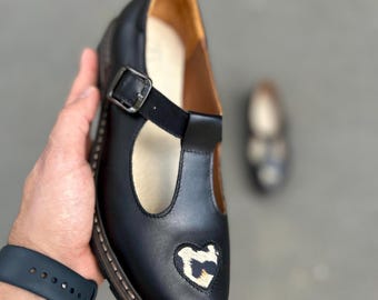 May include: Black leather Mary Jane shoe with a heart-shaped design on the toe. The shoe features a buckle closure, a T-strap, and a textured sole. The interior is a light brown color.