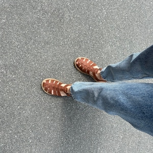 K&ouml;nnte beinhalten: Ein Paar braune Ledersandalen mit geflochtenem Design. Die Sandalen werden von einer Person getragen, die eine blaue Jeans tr&auml;gt.