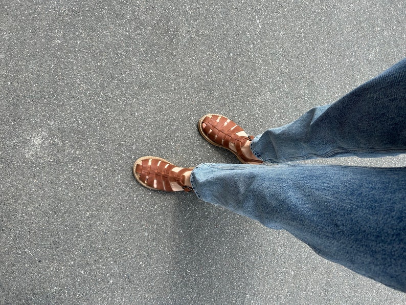 K&ouml;nnte beinhalten: Ein Paar braune Ledersandalen mit geflochtenem Design. Die Sandalen werden von einer Person getragen, die eine blaue Jeans tr&auml;gt.