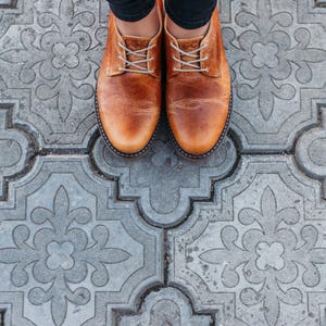 May include: A pair of brown leather shoes with laces. The shoes are on a gray patterned tile floor.