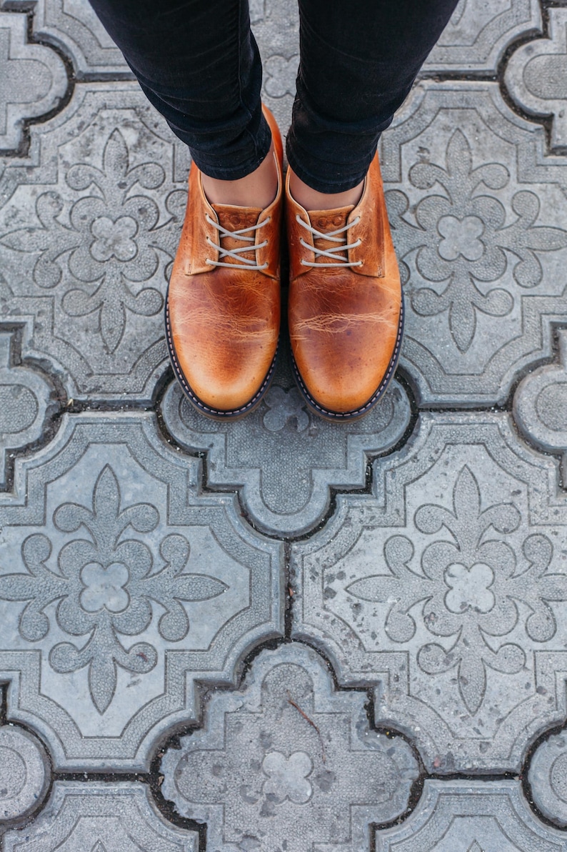 May include: A pair of brown leather shoes with laces. The shoes are on a gray patterned tile floor.