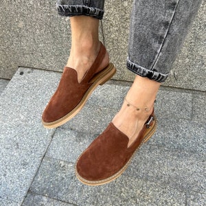 May include: Brown suede slip-on shoes with an ankle strap and a buckle closure. The shoes have a light brown sole and are worn by a person wearing jeans.