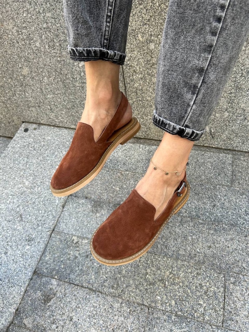 May include: Brown suede slip-on shoes with an ankle strap and a buckle closure. The shoes have a light brown sole and are worn by a person wearing jeans.