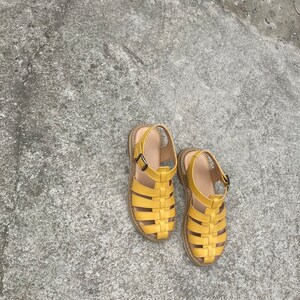 May include: A pair of yellow leather sandals with a woven design. The sandals have a buckle closure and a flat sole.