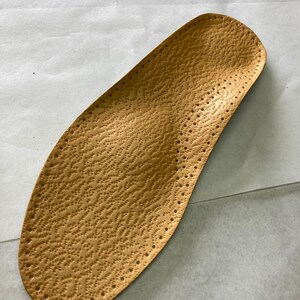May include: A beige leather insole with a patterned design and small holes throughout. The insole is designed to provide support and comfort for the feet.