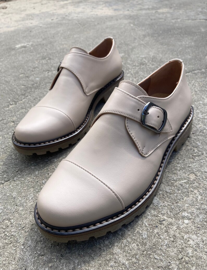K&ouml;nnte beinhalten: Ein Paar beige Lederschuhe mit Schnallenverschluss. Die Schuhe haben eine runde Spitze und eine dicke, strukturierte Sohle. Die Schuhe sind etwa 25 cm lang.