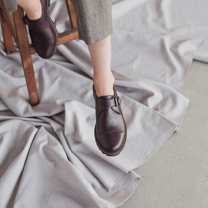K&ouml;nnte beinhalten: Ein Paar braune Lederschuhe mit einer Schnalle. Die Schuhe befinden sich auf einer wei&szlig;en Stoffoberfl&auml;che.