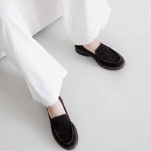 May include: A pair of brown suede loafers with a tassel detail. The loafers are worn by a person wearing white pants.