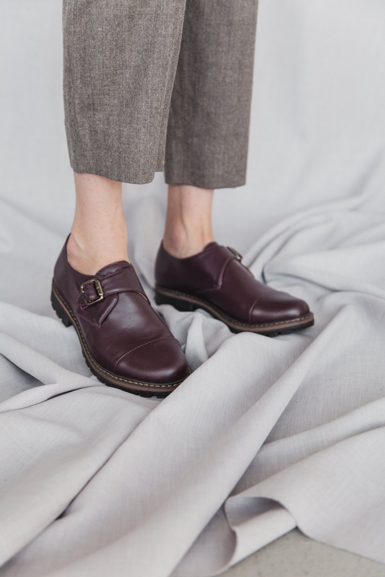 K&ouml;nnte beinhalten: Ein Paar burgunderfarbene Lederschnallen-Schuhe mit dicker Sohle. Die Schuhe befinden sich auf einer wei&szlig;en Stoffoberfl&auml;che.