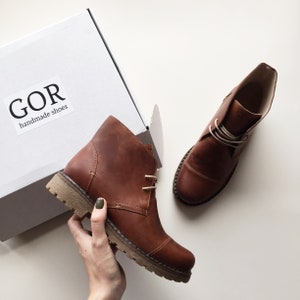 May include: A pair of brown leather ankle boots with white laces. The boots are handmade and the brand name "GOR handmade shoes" is printed on a white box.