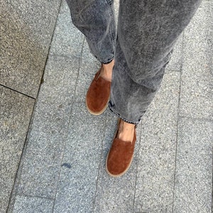 May include: A pair of brown suede loafers with a slightly pointed toe. The loafers are worn with dark gray denim jeans.