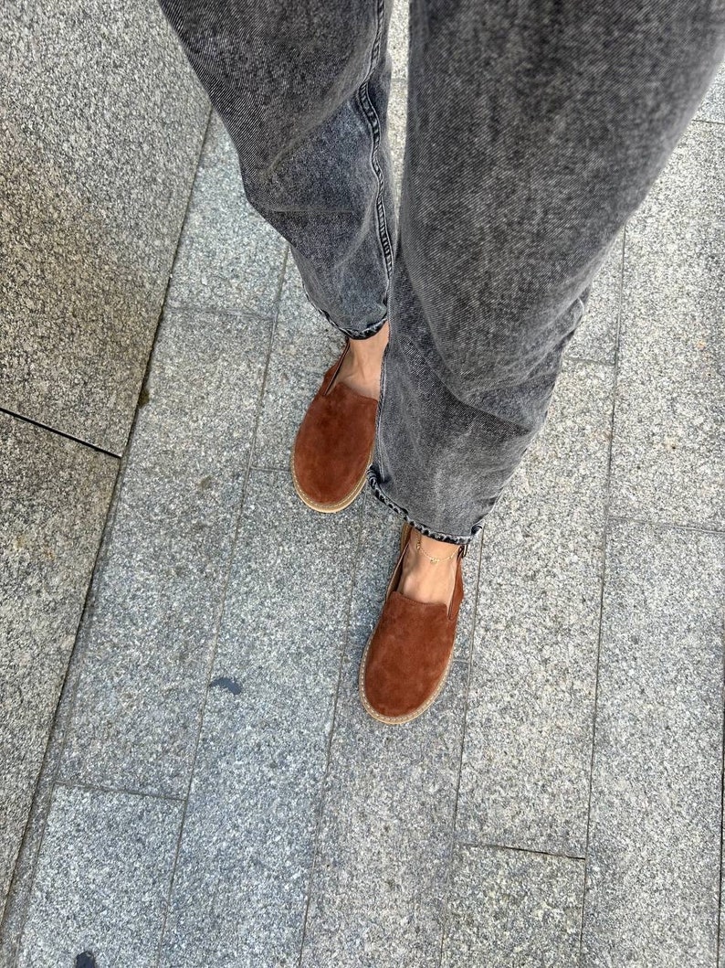 May include: A pair of brown suede loafers with a slightly pointed toe. The loafers are worn with dark gray denim jeans.
