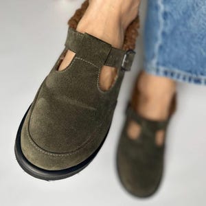 Pode incluir: Um par de chinelos de camurça verde com fecho de fivela e forro de pele castanho. Os chinelos estão a ser usados por uma pessoa.