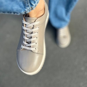 May include: A pair of gray leather low-top sneakers with white laces. The sneakers have a white sole and are worn with blue jeans.