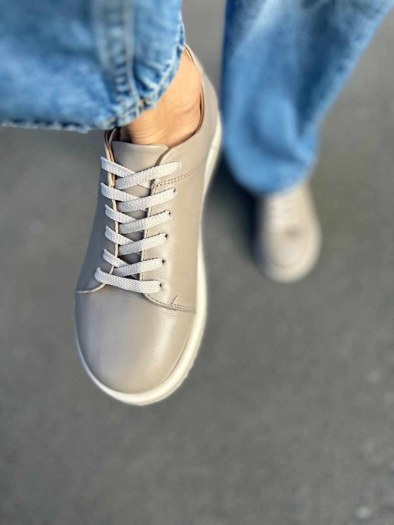 May include: A pair of gray leather low-top sneakers with white laces. The sneakers have a white sole and are worn with blue jeans.
