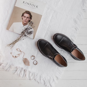 May include: Two black leather shoes with buckles on a white fuzzy surface. The shoes have a pointed toe and a thick sole. There is a magazine with the word "KINFOLK" on the cover and a bouquet of lavender on the surface.