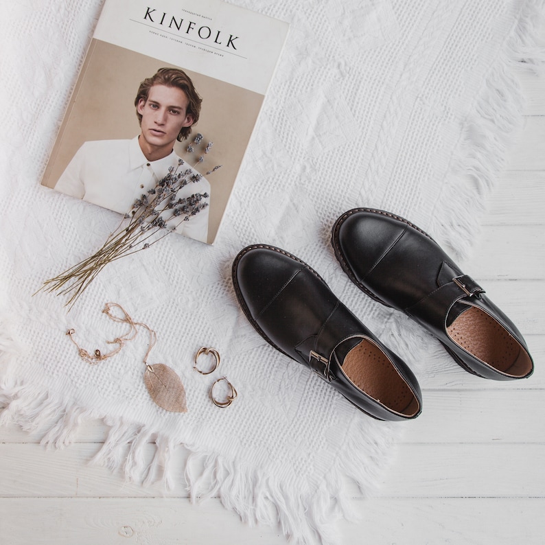 May include: Two black leather shoes with buckles on a white fuzzy surface. The shoes have a pointed toe and a thick sole. There is a magazine with the word "KINFOLK" on the cover and a bouquet of lavender on the surface.