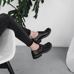 May include: A person wearing black pants and black leather shoes with buckles. The person is sitting in a white chair with black legs. A green plant is in the background.