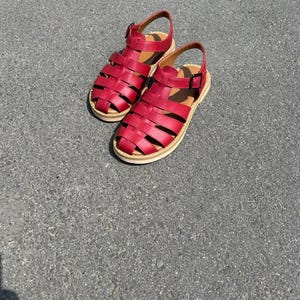 Puede incluir: Un par de sandalias de cuero rojo con dise&ntilde;o enjaulado. Las sandalias tienen cierre de hebilla y suela color beige. Los zapatos se muestran sobre una superficie gris.
