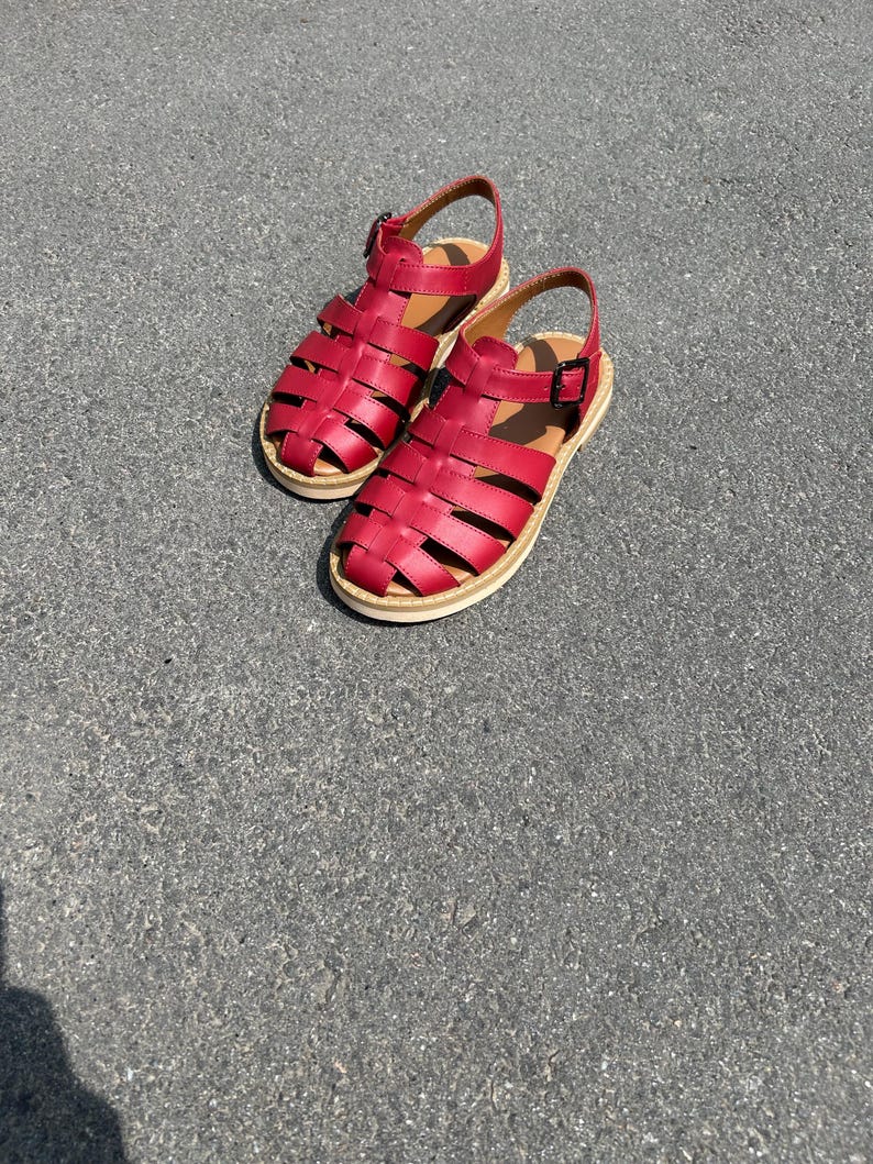 Puede incluir: Un par de sandalias de cuero rojo con dise&ntilde;o enjaulado. Las sandalias tienen cierre de hebilla y suela color beige. Los zapatos se muestran sobre una superficie gris.