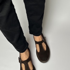 Puede incluir: Zapatos Mary Jane de ante marr&oacute;n oscuro con cierre de hebilla. Los zapatos tienen una puntera redondeada y un dise&ntilde;o de correa en T. Las suelas son de color marr&oacute;n claro. La persona lleva pantalones negros.