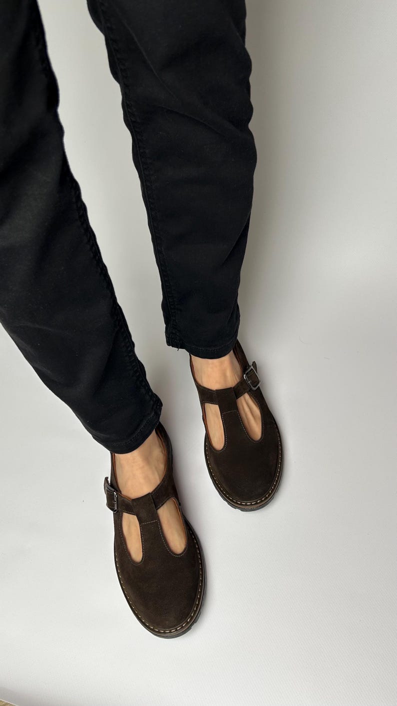 Puede incluir: Zapatos Mary Jane de ante marr&oacute;n oscuro con cierre de hebilla. Los zapatos tienen una puntera redondeada y un dise&ntilde;o de correa en T. Las suelas son de color marr&oacute;n claro. La persona lleva pantalones negros.