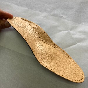 May include: A beige leather insole with a textured surface and small holes throughout. The insole is designed to provide support and comfort for the feet.