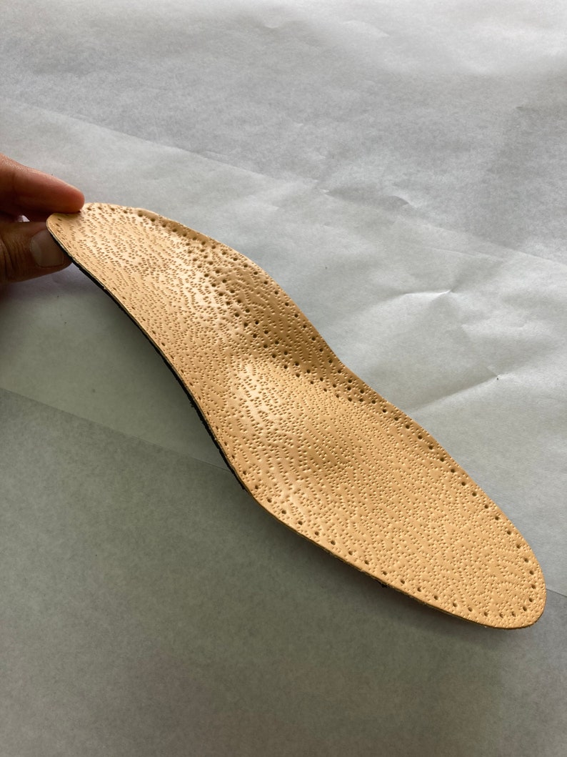May include: A beige leather insole with a textured surface and small holes throughout. The insole is designed to provide support and comfort for the feet.
