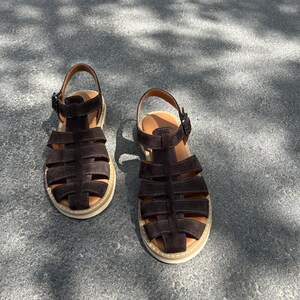 Pu&ograve; includere: Un paio di sandali in camoscio marrone scuro con un design a gabbia. I sandali hanno una suola color beige chiaro e una chiusura con fibbia. Le scarpe sono su una superficie di cemento grigio.