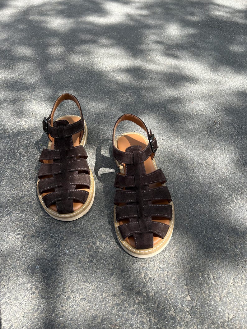 Pu&ograve; includere: Un paio di sandali in camoscio marrone scuro con un design a gabbia. I sandali hanno una suola color beige chiaro e una chiusura con fibbia. Le scarpe sono su una superficie di cemento grigio.