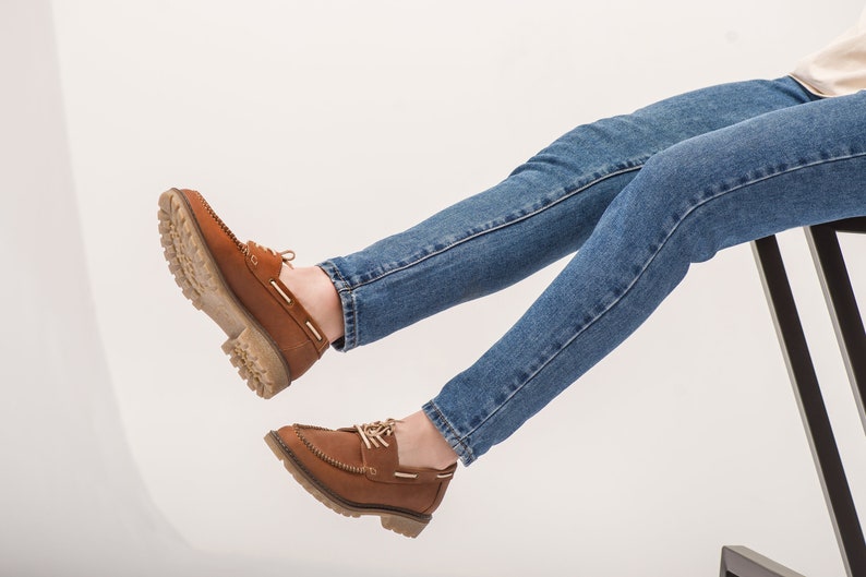 Op de afbeelding: Een paar bruine leren bootschoenen met witte veters. De schoenen worden gedragen door iemand die een blauwe jeans draagt.