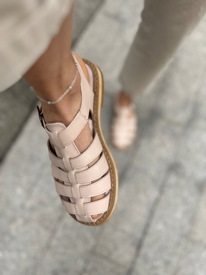 May include: A pair of light pink leather sandals with a caged design and a brown sole. The sandals have a buckle closure and are worn on a person's feet.