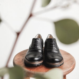 May include: A pair of black leather lace-up shoes with a brogue design. The shoes are sitting on a wooden stool.
