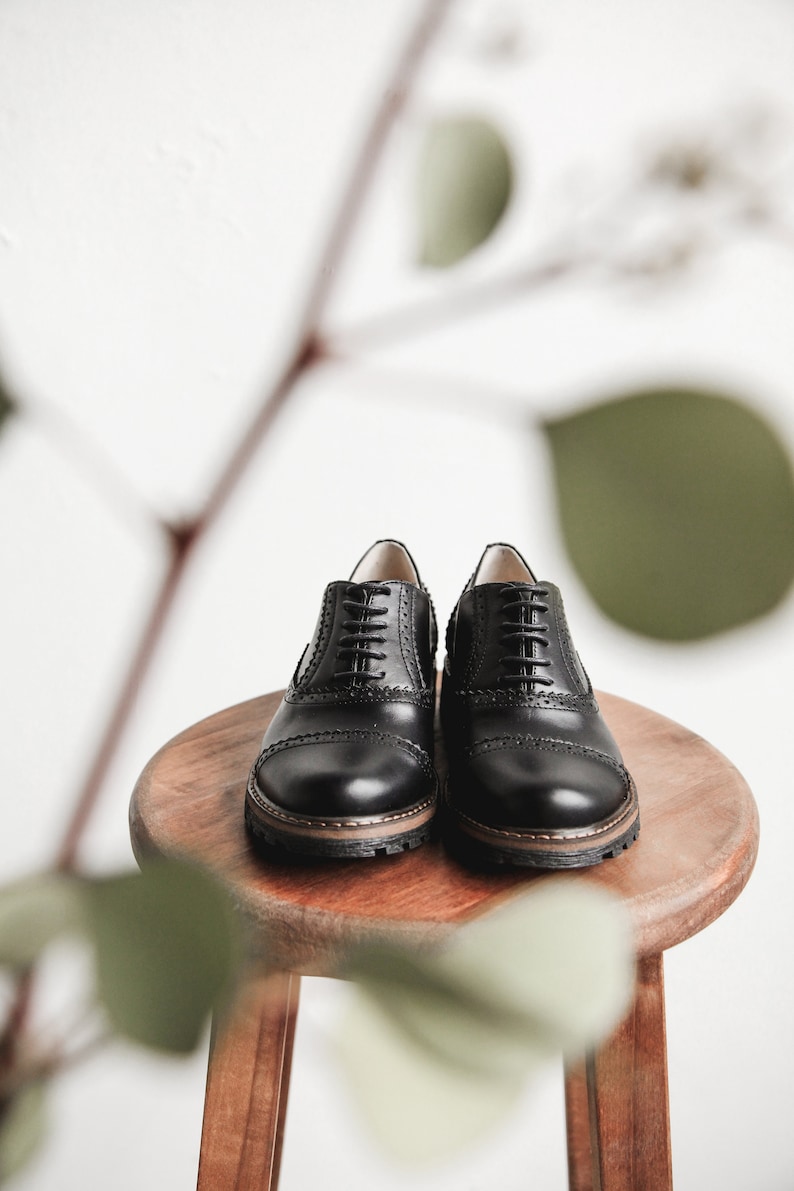 May include: A pair of black leather lace-up shoes with a brogue design. The shoes are sitting on a wooden stool.
