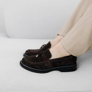 May include: Brown suede loafers with a decorative buckle on the front. The loafers have a black rubber sole.