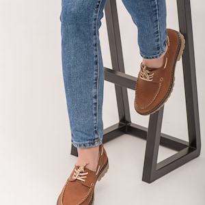 Op de afbeelding: Een paar bruine leren bootschoenen met veters. De schoenen worden gedragen door iemand die blauwe jeans draagt en op een zwarte metalen kruk zit.