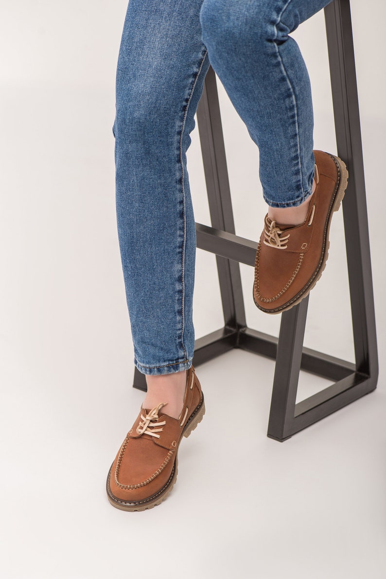 Op de afbeelding: Een paar bruine leren bootschoenen met veters. De schoenen worden gedragen door iemand die blauwe jeans draagt en op een zwarte metalen kruk zit.