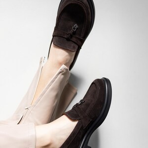 May include: A pair of brown suede loafers with a buckle detail. The loafers have a chunky black sole and are worn on a person's feet.