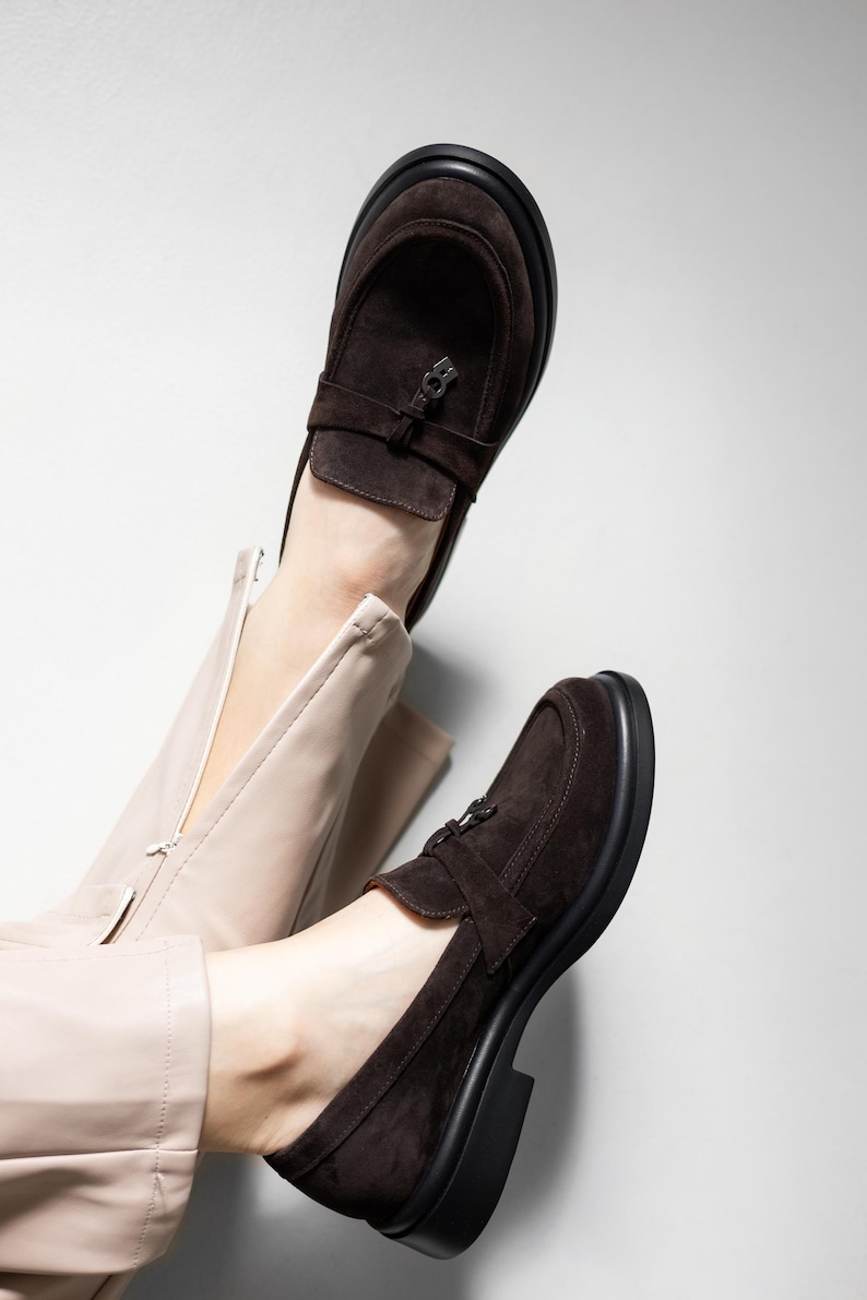 May include: A pair of brown suede loafers with a buckle detail. The loafers have a chunky black sole and are worn on a person's feet.