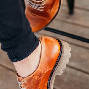 May include: A pair of brown leather lace-up shoes with a thick sole. The shoes are worn by a person wearing black jeans.