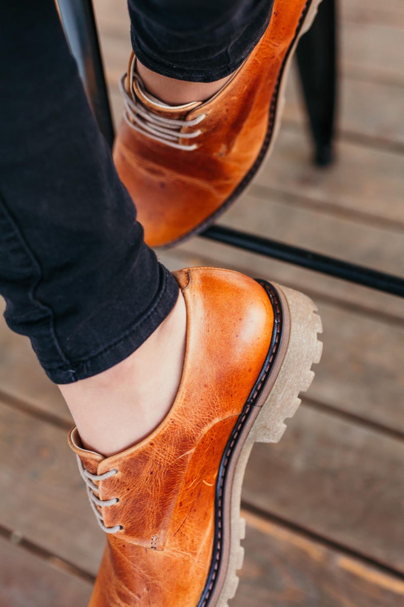 May include: A pair of brown leather lace-up shoes with a thick sole. The shoes are worn by a person wearing black jeans.