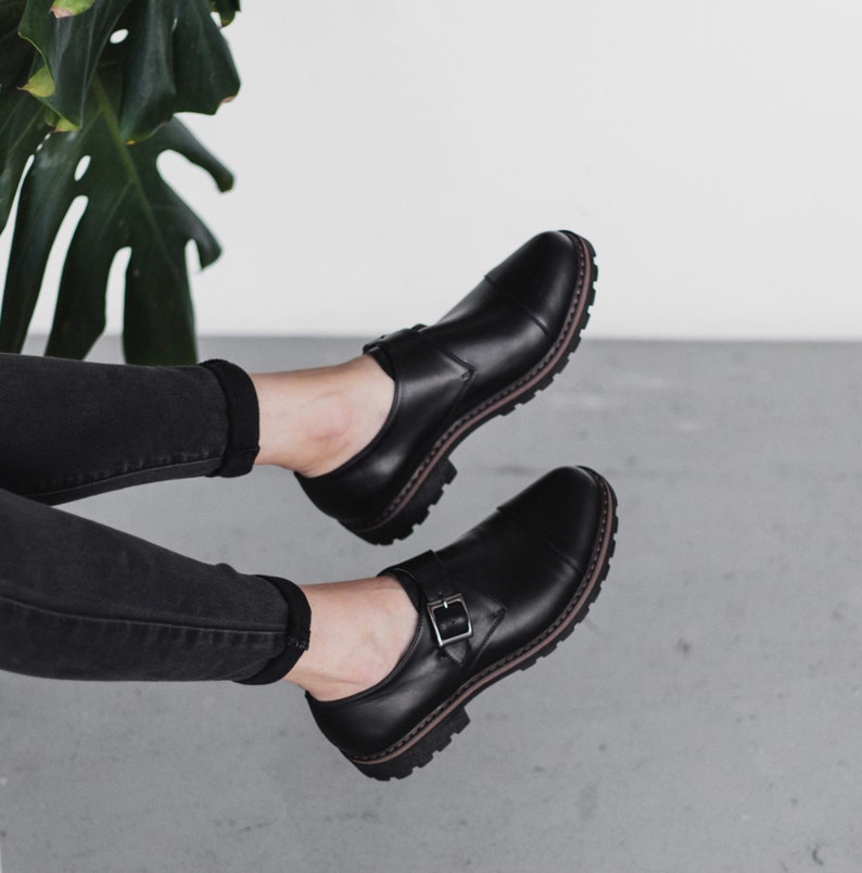 May include: Black leather buckle shoes with a chunky sole. The shoes are worn by a person with black jeans.