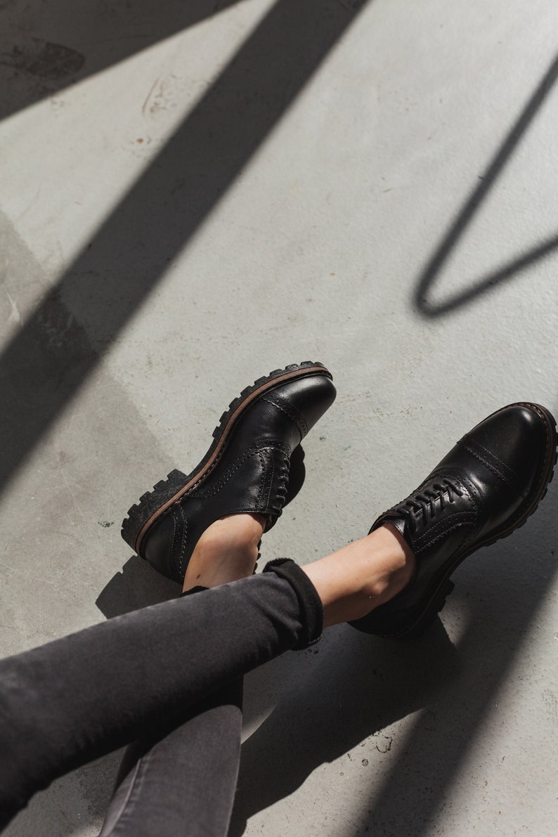 May include: A pair of black leather lace-up shoes with a chunky sole. The shoes are worn by a person sitting on a concrete floor with their legs crossed.