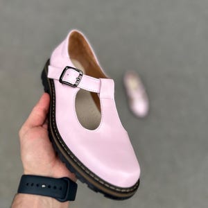 Peut inclure: Une chaussure Mary Jane rose clair avec une boucle noire et une semelle épaisse bordée de marron. La chaussure est tenue dans une main, mettant en valeur son design et sa couleur. La semelle semble faire environ 2,5 cm d'épaisseur.