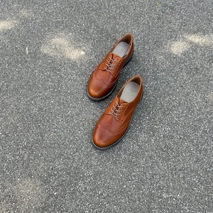 Pu&ograve; includere: Un paio di scarpe brogue in pelle marrone dal design classico. Le scarpe hanno uno stile wingtip con perforazioni decorative e chiusura con lacci. Le scarpe sono appoggiate su una superficie grigia.