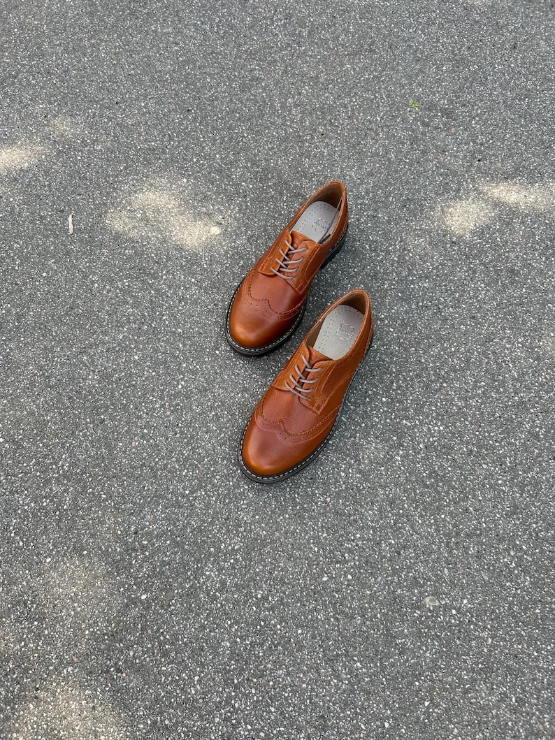 Pu&ograve; includere: Un paio di scarpe brogue in pelle marrone dal design classico. Le scarpe hanno uno stile wingtip con perforazioni decorative e chiusura con lacci. Le scarpe sono appoggiate su una superficie grigia.