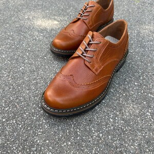 Pu&ograve; includere: Un paio di scarpe Oxford in pelle marrone con perforazioni decorative e lacci colorati chiari. Le scarpe hanno un classico design a punta alata e una suola robusta.